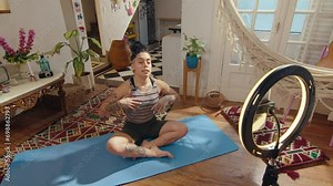 Young yoga instructor sitting cross legged on mat, talking about practice and showing side bend exercise in front of phone with ring light while filming stretch class for fitness channel