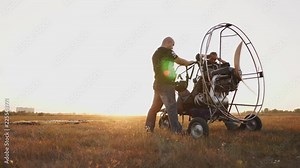 The motor paraglider stands in a field at sunset with a wooden propeller, two pilots conduct a test run of the engine before the flight. The camera moves dynamically on the gimbal