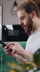 Cheerful man sitting on sofa uses mobile phone smiles at home. Guy texting message scrolling tapping technology isolated lifestyle. Slow motion.