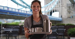 Pretty waitress carrying tray with coffee near Tower Bridge, Friendly young woman in England wearing apron and holding tray to serve coffee, 4k