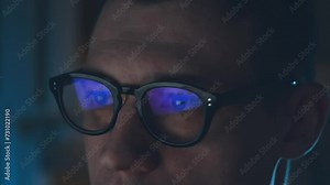Handsome man in computer glasses sits at a computer in the dark and types in code.