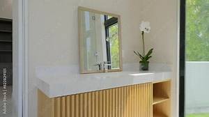 Elegant bathroom vanity area with clear view of rectangular mirror, wooden countertop and vase with white orchid. Slat design and greenery outside window