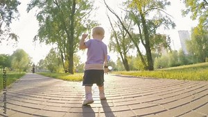 Baby girl one year old makes the first steps in a summer park, backlight, low wide angle shooting, slow motion