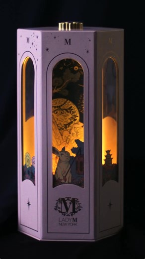 LADY M 流轉月光中秋月餅禮盒，以柔和粉色搭配細緻燙金，盒身典雅別緻。禮盒內建開關，開啟後燈光隨即亮起，緩緩自動旋轉，生動呈現玉兔穿越森林、奔月的奇幻之旅。採用 USB 充電設計，便利環保，隨時點亮團圓夜晚。 禮盒內附有典雅提袋與專屬賀卡，讓心意流轉其中，為美好的中秋時節增添溫度與儀式感。 | Lady M Taiwan