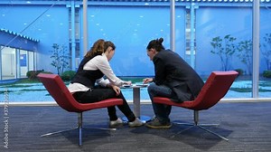 Man and woman lean over small table, using phones. Passengers sit on inclined chairs at domestic terminal lounge at modern Japanese airport, glass wall on background