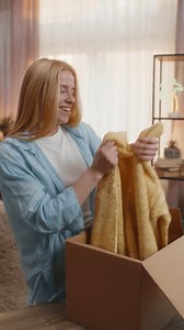 Young woman opens delivery box at home, removing taking blouse sweater clothing with excited grin and showing to camera. Caucasian girl feels satisfaction, enjoying new arrivals under evening sunset.