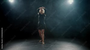Graceful blonde woman in black dress dancing alone in dark studio, slow motion. Beautiful lady performing contemporary choreography on stage, rehearsing in dance hall, bright soffit and artistic smoke
