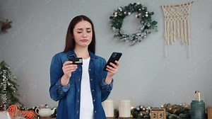 Woman stands in Christmas kitchen hold credit card use phone try to pay through e-bank app experiences problems no money, insecure online payment, scam, fraud, unsuccessful transaction error purchase.