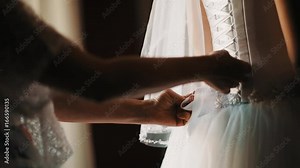 Female hands tights up young bride white wedding dress corset in semi dark room in front of window