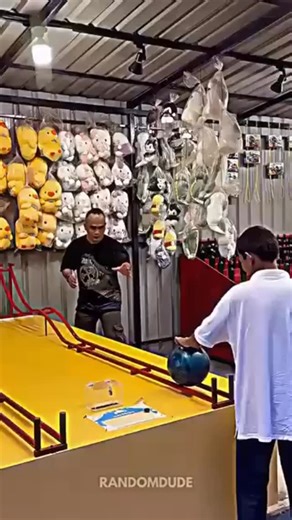 ExplainReel.io™ on Instagram: "- The post features a viral video of the “Bowler Roller” carnival game, where multiple players fail to roll a bowling ball up a yellow ramp into a distant hoop, but the booth operator nails it on the first try. - The operator’s success uses backspin physics: reverse rotation imparts angular momentum that, through ramp friction, slows the ball precisely at the hoop, as confirmed by similar viral clips on TikTok and Instagram. - Replies highlight the game’s deceptive