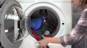 A young woman is actively loading clothes with a washing machine. The woman opens the washing machine and throws clothes into her then closes it.