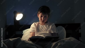 Boy resting in his bed in the evening and reading a book turns off light and goes to sleep