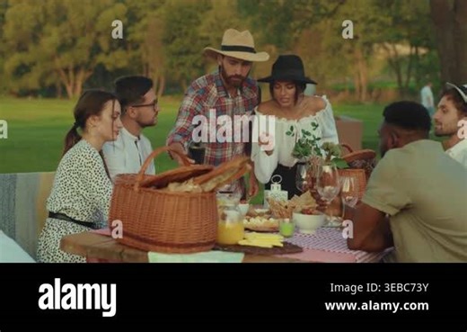 Picnic table set with wine glasses and wicker basket, host pours bottle while friends sample cheese and bread, warm Stock Video Footage - Alamy