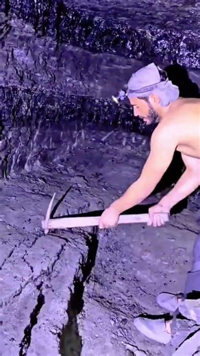 digging dark minerals in a cave using a pickaxe while wearing a headlamp to illuminate the workspace