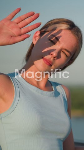 Young lady resting right hand on iron railing, using left hand to block sunlight from eyes, squinting under bright sun, background blurred with distant greenery | Premium Stock Video Footage