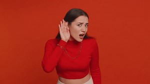 I cant hear you. What. Young woman trying hear you, looking confused and frowning, keeping arm near ear for louder voice, asking to repeat to hear information, deafness. Girl on red studio background
