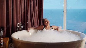 Asian senior woman relax in warm bubble bath. Old lady enjoys in freestanding bathtub full of soap foam. Mature biethnic female wash body at luxury spa hotel bathroom tub, panoramic city and sea view.