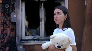 Young sad woman with teddy bear standing on the right at background of bombed house sighing looking away. Thoughtful Ukrainian beautiful lady with toy outdoors at ruined property