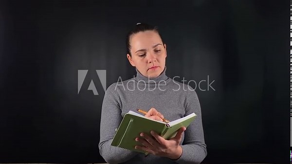 A portrait of a beautiful young white woman holding a notepad, opening it, jotting down notes, making entries in a planner on a black background. Planning and record-keeping in a purposeful lifestyle