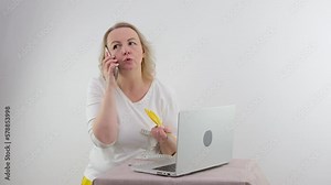 middle-aged woman on white background is standing Near a table with a laptop hands in sideways looks into the frame smiling holding her lips incomprehension surprise expectation space for text