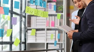 Professionals brainstorming and discussing ideas with sticky notes on a glass board. Collaborative team planning and analyzing project details in an office.