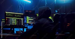 Back view on the male software developer in hood working over hacking programs at the computer with codes on the screen at night in the dark room. Rear.