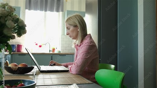 Middle-aged blonde businesswoman typing on a laptop keyboard and using a touchpad while sitting at a kitchen table. Professional freelancer concentrating on a project in a home office environment