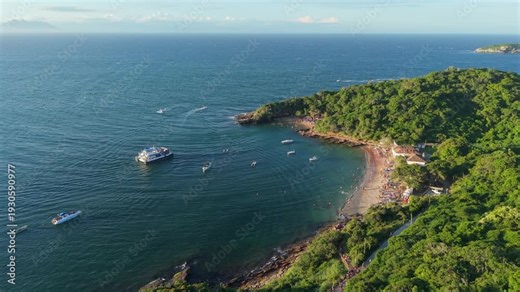 Bustling Praia da Azeda in Armação dos Búzios, Rio de Janeiro, Brazil, forming sheltered turquoise cove along South Atlantic shoreline with Mata Atlântica hillside and anchored boat, drone gliding