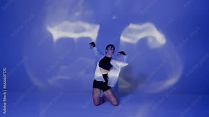 Young woman wearing a top, shorts and a shirt performing contemporary dance under the glare of light in the studio. Neon blue color scheme, shadowed background. Full length.