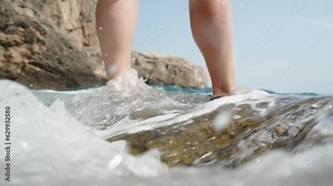 Low-angle shot capturing the view of female feet being covered by the waves of the sea as she stands on a rock at the shore, in slow motion.