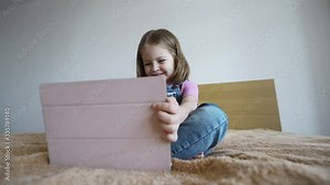 From below angle view of a laughing blonde little girl holding a pink tablet in her hand, sitting on a bed, close-up