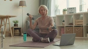 Full shot of strong mature female gym coach working remotely providing exercise example to client through video call sitting in butterfly pose lifting small dumbbells