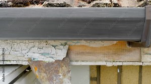 Closeup POV elevated outside shot at house roof level, of a hand using a scraper to remove peeling white gloss paint on a wooden fascia board, beneath rain guttering.