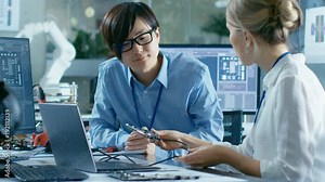 Two Computer Engineers Talking, They Connect Printed Circuit Board to a Laptop and Start Coding. In the Background Computer Science Research Laboratory with Robotic Arm Model.