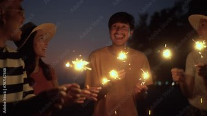 Happy friends group partying on the beach dancing and waving sparklers on summer at night. Group people having fun and enjoy on beach party smiling and dancing with sparklers.