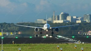 Airliner taking off from the runway in a front view with both wings visible. The aircraft is gaining speed for ascent, with a blurred cityscape in the background under a clear sky