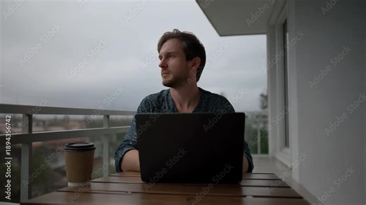 Man Using Laptop on Balcony Table Amidst Cloudy Sky, Facing Camera, Coffee Cup Nearby, Relaxed Mood today