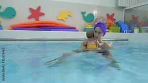 A young female instructor is engaged in developmental exercises with a newborn baby in the water in the children's pool. She holds him in her arms and hugs him. Infant swimming. Closeup.