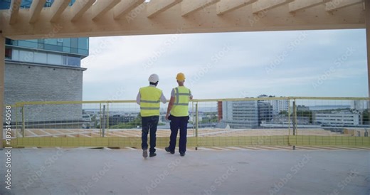 Two Male Construction Engineers Walk Across Building Construction Site, Discussing Structural Details, Commercial and Residential Potential, Reviewing the Modern Real Estate Development Project