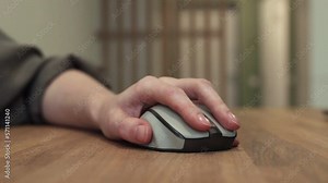 Hand on computer mouse, finger clicking key, pressing left button and moving device on wooden desk closeup