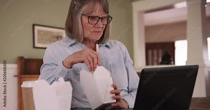 Retired senior woman having lunch and using laptop in domestic home setting, Caucasian lady eating while watching something on portable computer, 4k