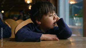 Bored small boy yawning while waiting at restaurant table by window. Child feeling boredom in authentic lifestyle moment awaiting for food to arrive