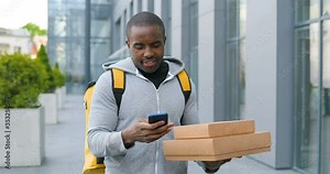 African American deliveryman with carton parcels standing the street, tapping on navigation map on phone looking for address. Male courier using navigating system on smartphone to deliver boxes.