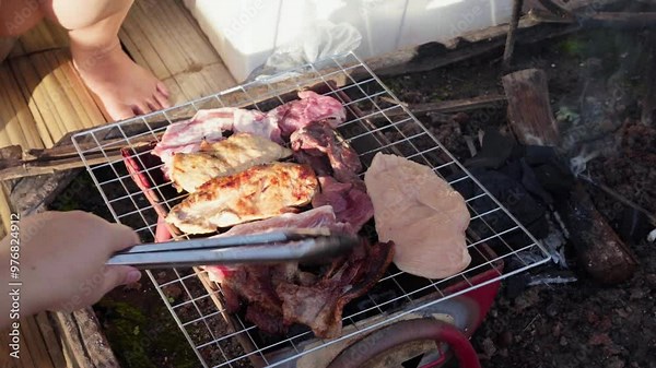 A close-up 4K HD cinematic shot of a barbecue grill with wood and charcoal ember burning. The scene shows someone turning chicken and pork on the grill with metal tongs, sizzle and cooking process
