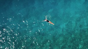 Woman swimming in clear ocean waters. Aerial top down view of adult girl relaxing alone in the vast, blue sea.