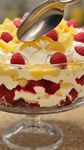 Lemon Raspberry Trifle of Golden Layers 🍋 Pale lemon yellow cream layers alternate with vivid ruby raspberry compote inside a clear glass trifle bowl. The textures are distinctly stratified: airy whipped cream with soft peaks, silky lemon curd ribbons, tender sponge cake soaked lightly, and juicy whole raspberries embedded between layers. The top is lavishly crowned with billowy whipped cream, fresh raspberries dusted lightly with sugar, and thin lemon slices curled like petals. Colors are clea