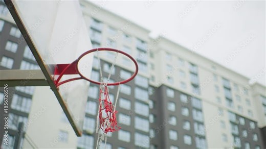 urban basketball hoop city building backdrop, ball arcs toward rim then empties net, red metal rim with frayed chain net, overcast sky, residential highrise windows blurred, quiet neighborhood court,