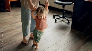 Little feet walking on floor, close-up. Baby learning to walk at home. Baby first steps. Smiling caring mother and cute little infant child girl cuddling in bedroom. Mum and child tender moments.