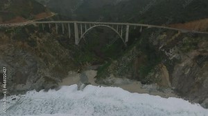 Aerial footage of Bixby Creek Bridge spanning valley at sea coast. Waves rolling on sand. Hazy view of mountain landscape in background. California, USA
