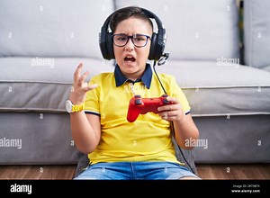 Young hispanic kid playing video game holding controller wearing headphones crazy and mad shouting and yelling with aggressive expression and arms rai Stock Photo - Alamy